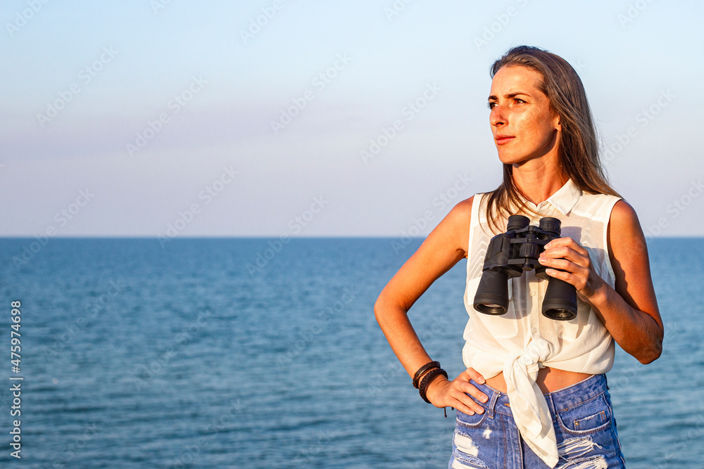 Beautiful young woman with binoculars on the cliff looks at the sea.
