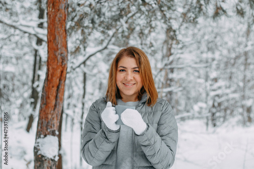 A cute girl with short hair in a gray jacket and knitted white mittens is drinking hot tea in a snowy forest. Cozy and warm in the winter season.selective focus