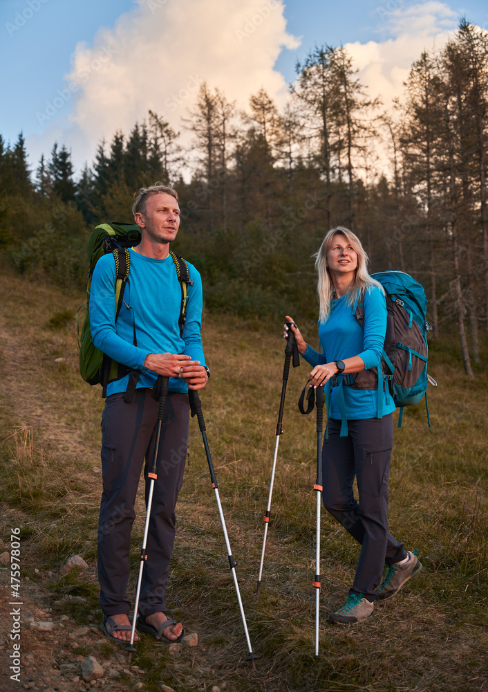 Two travelers with trekking poles during hike, stopping on mountain trail and admiring the surrounding natural scenery. People going adventural trek in the mountains.