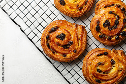 Just baked pain aux raisins on cooking wire rack. Buns are also called escargot or pain russe, is a spiral pastry with custard cream and raisin. Directly above, white table surface. photo
