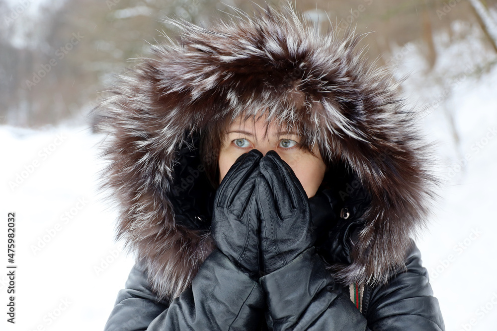 Woman in leather coat with fur hood standing on a street during snow and covering her face by hands in gloves. Cold weather in winter city Woman in leather coat with fur hood standing on a street during snow and covering her face by hands in gloves. Cold weather in winter city