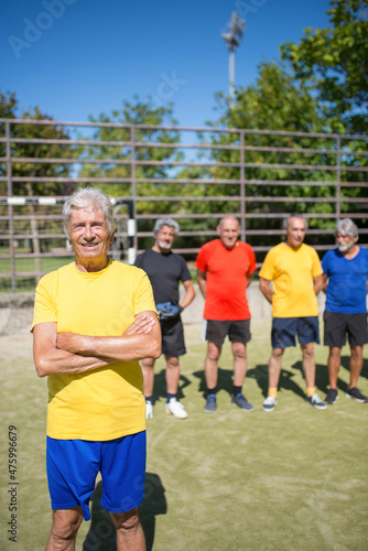 Portrait of content senior man on football field. Captain with gray hair in sport clothes standing, looking at camera, teammates in background. Football, sport, leisure concept