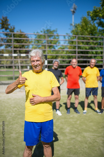 Portrait of contented senior man on football field. Captain with gray hair in sport clothes standing, looking at camera, teammates in background, showing thumb up. Football, sport, leisure concept