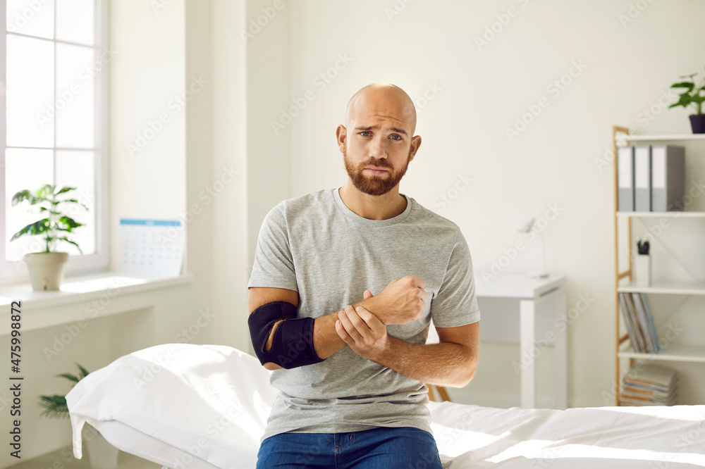Man wears black elastic supportive bandage on his elbow. Portrait of sad man who is forced to wear bandage on his arm after bone fracture. Man sits on couch in doctor's office and looks at camera.