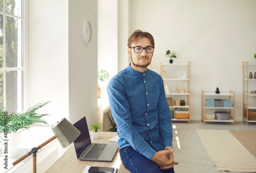 Portrait of professional young male manager near his workplace in modern office. Handsome Caucasian man in glasses, jeans and shirt stands leaning on his desk and looks at camera.