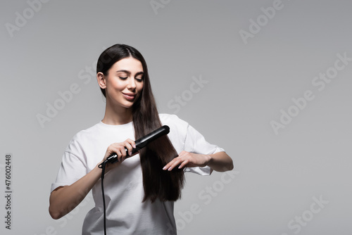 positive young woman using hair straightener isolated on grey photo