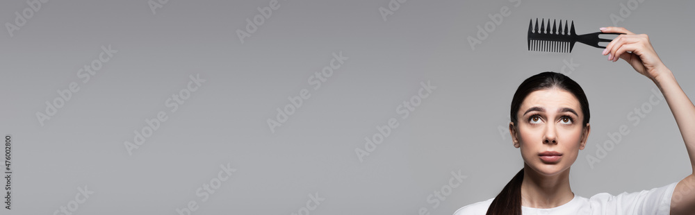 Naklejka premium worried woman in white t-shirt holding comb above straight hair isolated on grey, banner
