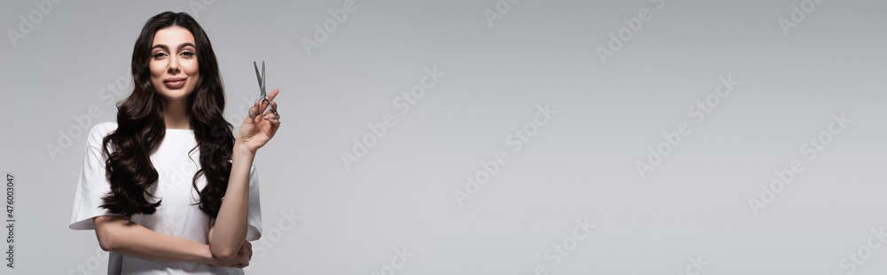 positive young woman with long wavy hair holding scissors isolated on grey, banner