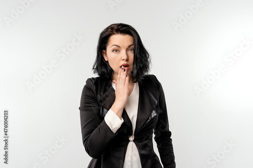 Coy cute young businesswoman in black formal jacket white shirt covering mouth and looking guilty, did something awkward, standing over white background