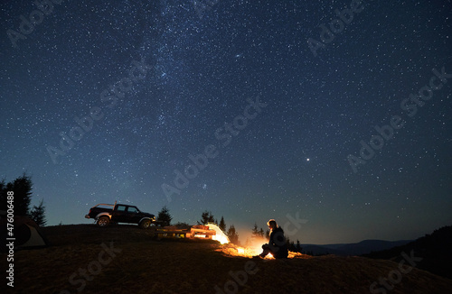 Female camper resting by bonfire and enjoying pleasant atmosphere in camp outdoor. Two tents  wooden table between them and black jeep parked on hill. Beautiful shining stars in the night sky.