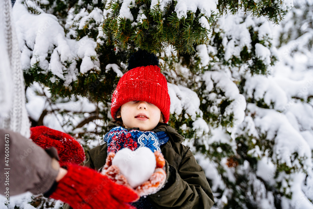 Unrecognizable mother giving snowball shape heart for her little son, dressed in colorful clothes are playing together with snowball in a winter park with snowy spruce on background. Unrecognizable mother giving snowball shape heart for her little son, dressed in colorful clothes are playing together with snowball in a winter park with snowy spruce on background.