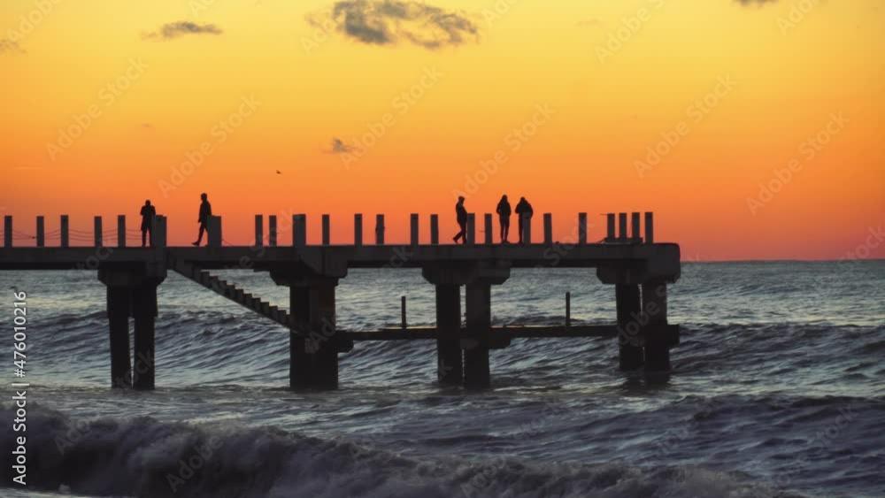 hot storm waves hit pier at sunset. Beautiful orange sky, crashing waves. Seascape. Resort romantic view, silhouettes of unrecognizable people walking along pier and enjoying scenery. hot storm waves hit pier at sunset. Beautiful orange sky, crashing waves. Seascape. Resort romantic view, silhouettes of unrecognizable people walking along pier and enjoying scenery.