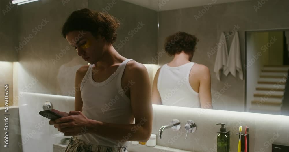 Young man with patches is typing text on a mobile phone standing in a bathroom Young man with patches is typing text on a mobile phone standing in a bathroom