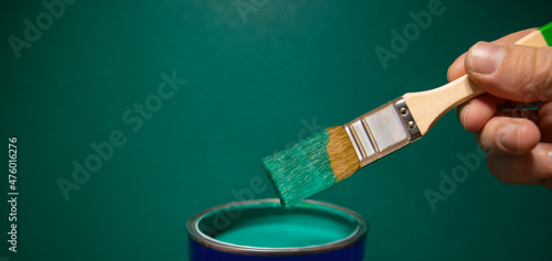 Man's hand holding a paintbrush over a can of paint on a green wall background, empty space for your text, banner, copyspace, advertisement or your design.