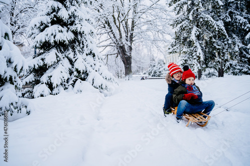 Two different age brothers kids have fun in the beautiful winter park with snow-covered trees. Children walk along a snowy road riding on sled. Winter bright knitted and warm clothes.