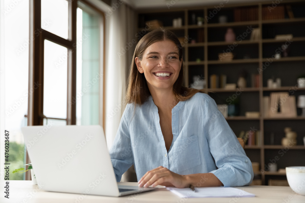 Cheerful young female worker sitting at workplace with laptop in office, looking away, smiling, laughing. Millennial employee, business woman in casual distracting from work, talking to colleagues