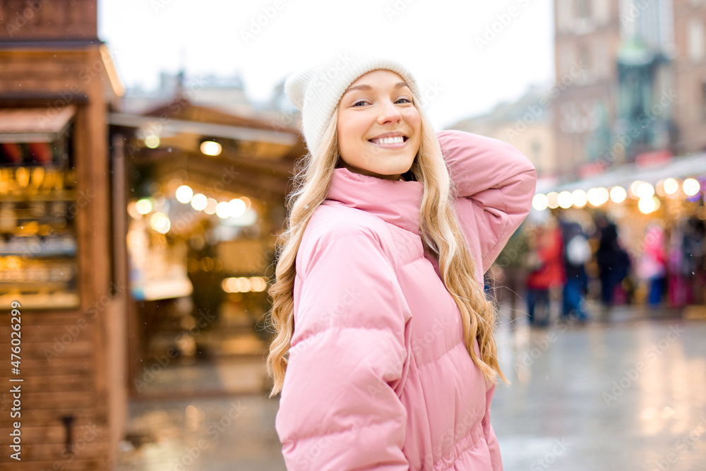 A cute blonde in a hat and a pink jacket walks around the city, winter fairs? Christmas mood