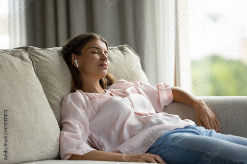 Relaxed calm girl resting, drowsing on couch, sleeping with wireless earphones on ear, listening to chill out, stress relief, recreation music, meditation audio course. Young woman taking break photo