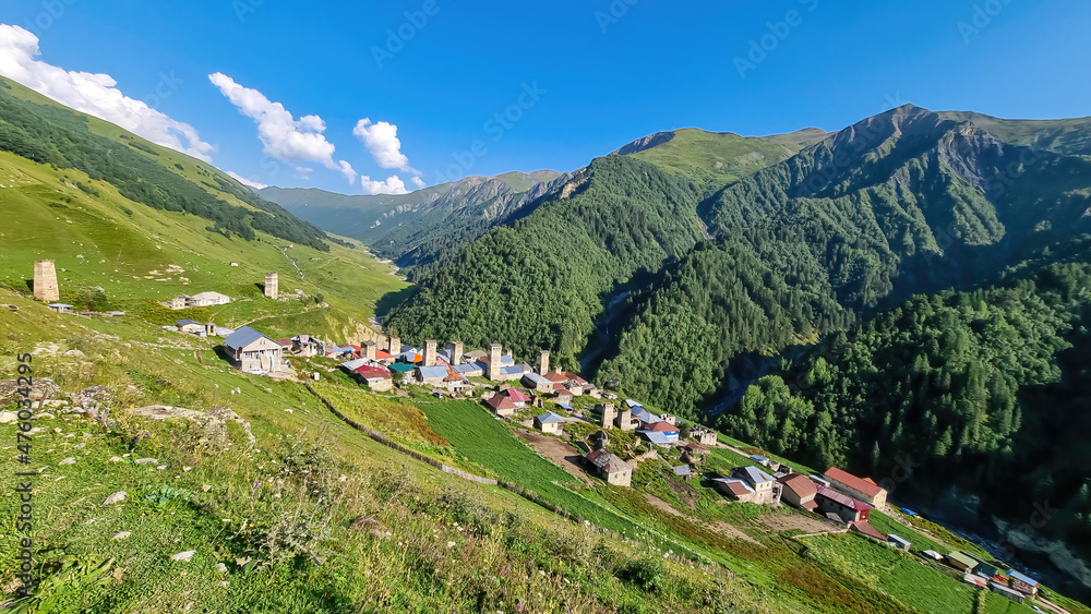 Panoramic view on Adishi, a mountain village, located in high Caucasus mountain chains. Few building on the bottom of the valley. Lush green pastures with a few cows grazing on them. Idyllic landscape