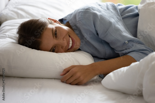 Happy beautiful young woman resting in bed, lying in white linen with head on comfortable pillow, looking at camera with toothy smile. Slumber girl enjoying awaking in morning after sleep enough