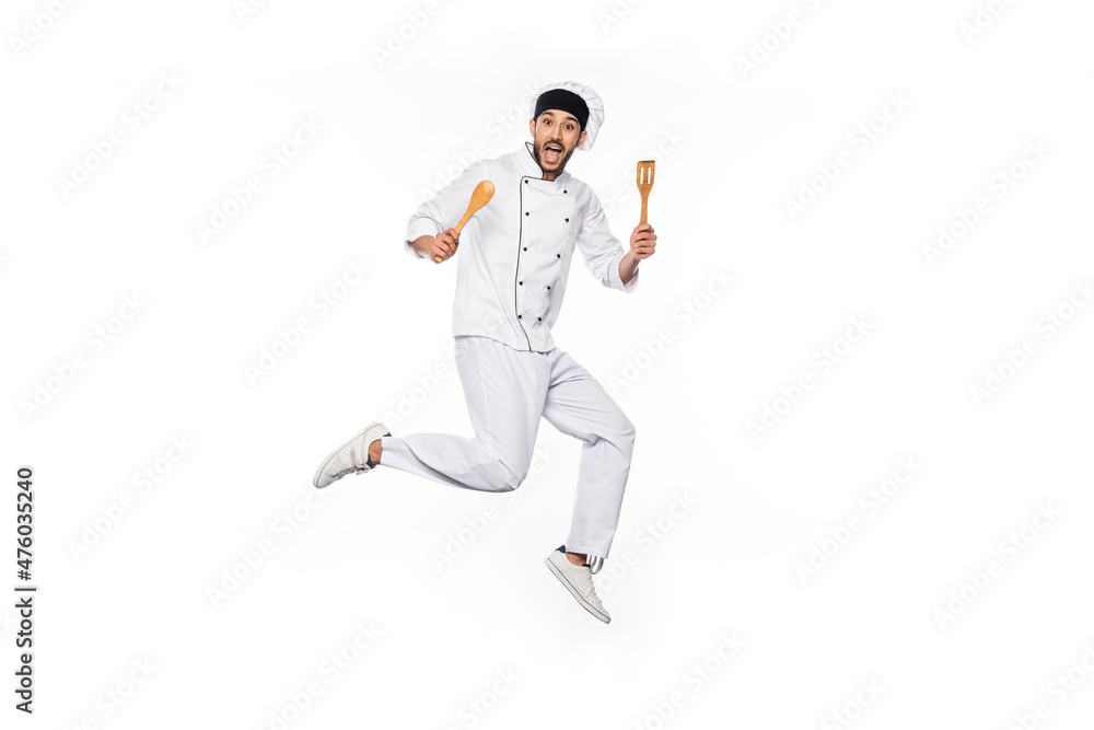 excited chef in hat and uniform jumping and holding wooden spatula and spoon isolated on white. excited chef in hat and uniform jumping and holding wooden spatula and spoon isolated on white.