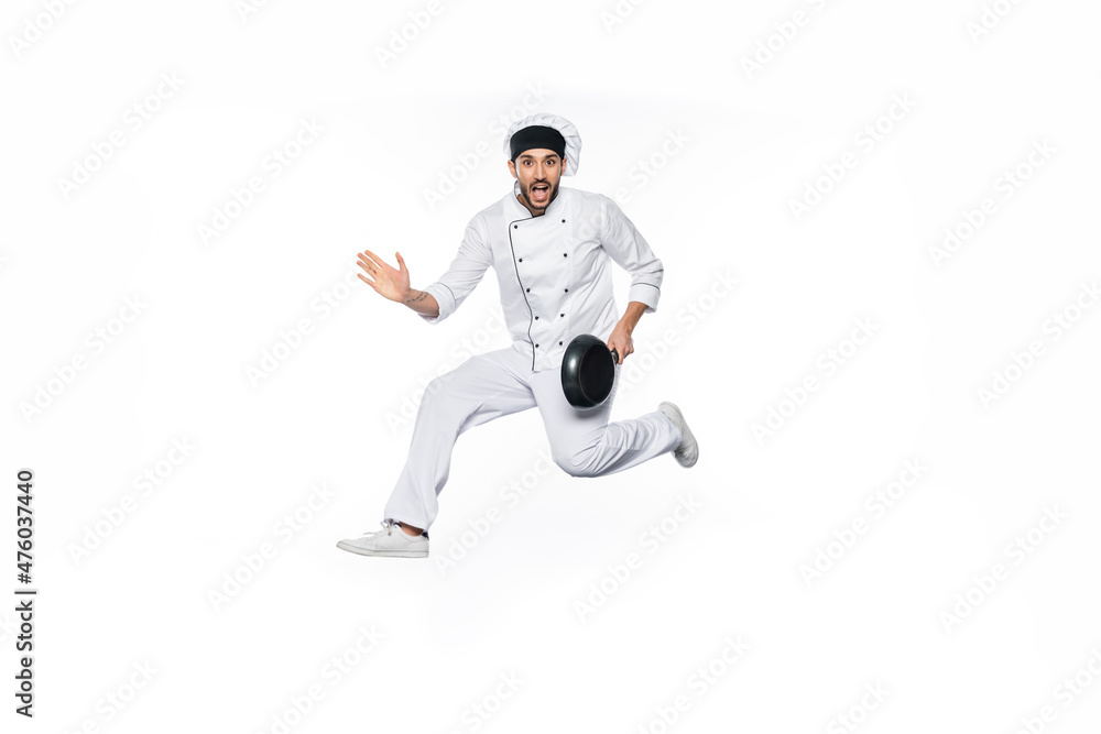 amazed chef in hat and uniform jumping and holding frying pan isolated on white.