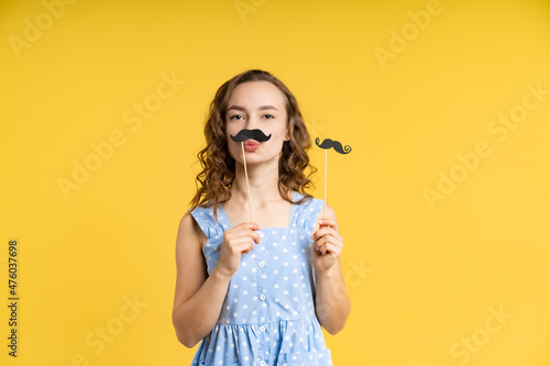 portrait of a beautiful girl in the studio. a model in a blue dress with white polka dots, with a mustache on a stick, jokes, fools around, has fun. yellow background.