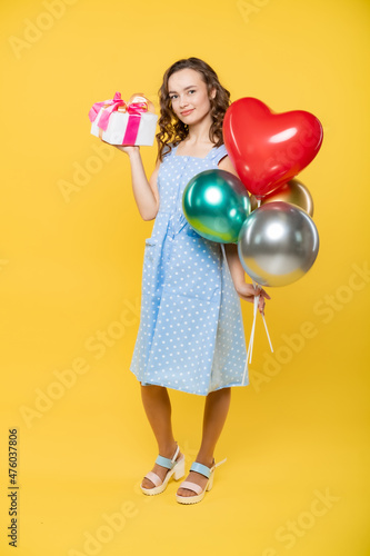 portrait of a beautiful girl in the studio with balloons as a heart. valentine's day, holiday, birthday. model in a blue dress with white polka dots. yellow background. holiday, birthday