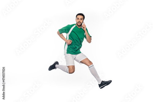 happy football player in uniform jumping while talking on smartphone isolated on white.