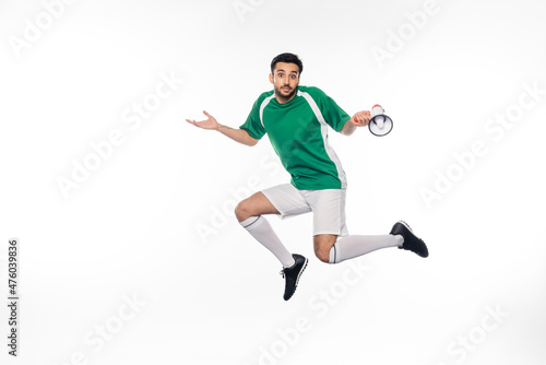 confused football player in uniform jumping and holding loudspeaker on white.