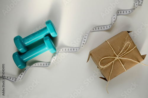 Two sports dumbbells, gift box and white measuring tape on white background with copy space. Christmas sport flatlay. Home training, gift.