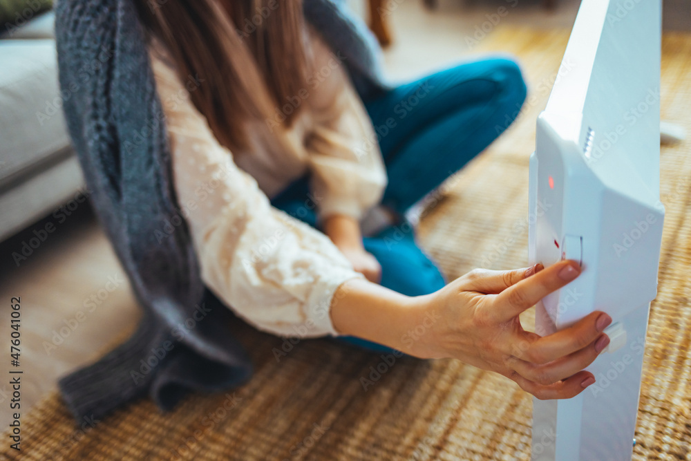 Fototapeta premium Using heater at home in winter. Woman regulating temperature on heater. Heating season. Young woman warming her hands on a heater. Smiling woman warms hands near radiator at home