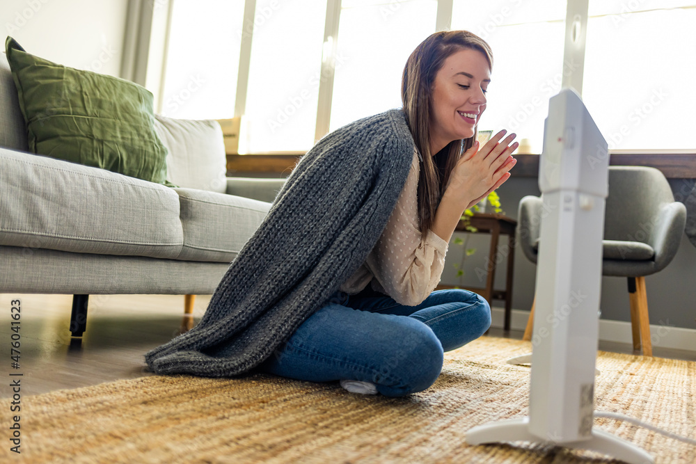 Smiling woman warms hands near radiator at home. Using heater at home in winter. Woman warming her hands. Heating season. Young woman sitting in room with electric heater. Smiling woman warms hands near radiator at home. Using heater at home in winter. Woman warming her hands. Heating season. Young woman sitting in room with electric heater.