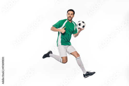amazed football player in uniform jumping while holding soccer ball and showing thumb up on white. © LIGHTFIELD STUDIOS