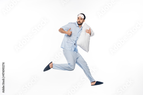 bearded man in pajamas and sleeping mask levitating with pillow while showing thumb up on white. © LIGHTFIELD STUDIOS