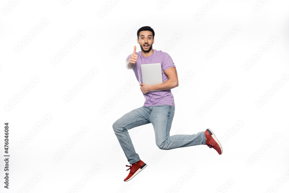 positive man in jeans and purple t-shirt levitating while holding laptop and showing thumb up on white. positive man in jeans and purple t-shirt levitating while holding laptop and showing thumb up on white.