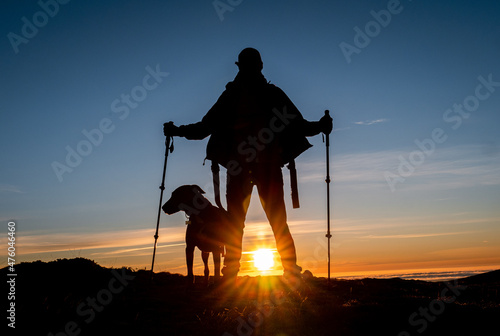 Caucasian male with hicking sticks and a dog standing on the top of a mountain at the sunset photo
