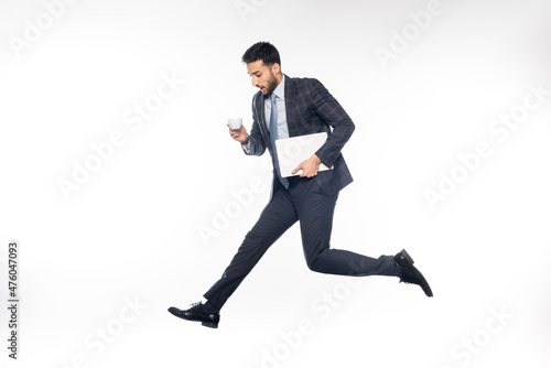 shocked businessman in suit jumping while holding laptop and cup on white.