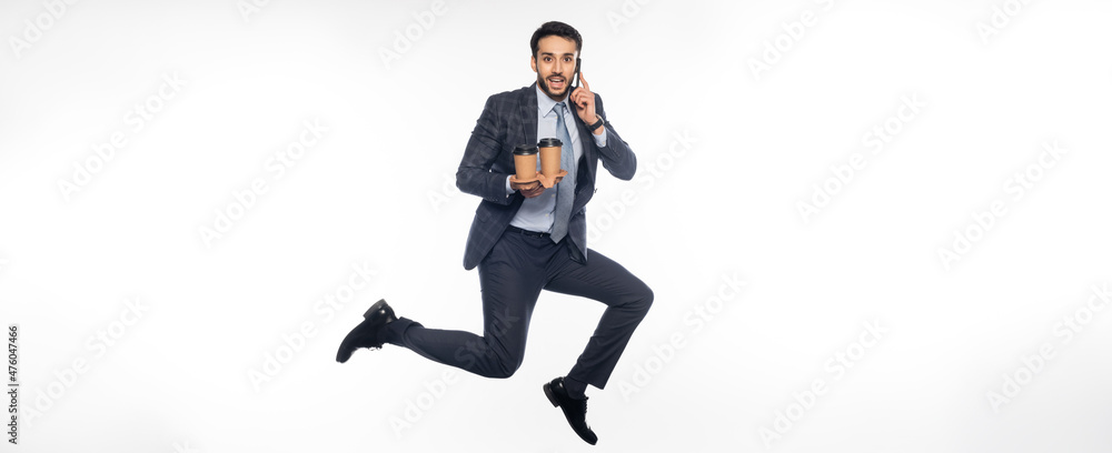 cheerful businessman in suit jumping while holding cup holder with paper cups and talking on smartphone on white, banner.