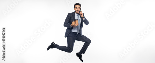 cheerful businessman in suit jumping while holding cup holder with paper cups and talking on smartphone on white, banner.
