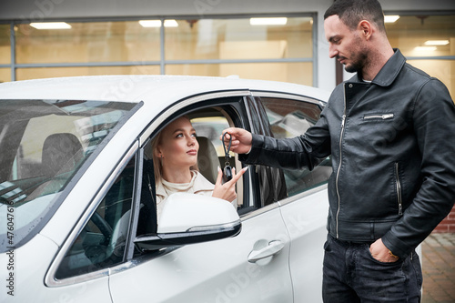 Handsome salesperson giving keys to young beautiful woman customer sitting in white car. Happy girl receiving key of her new automobile at auto dealership outdoors. © anatoliy_gleb