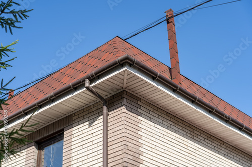 Brown roof of house covered with flexible bitumen motleysoft shingles under blue sky on sunny day. Cottage roof with metallic guttering system, guttering and drainage pipe exterior