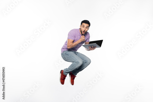 shocked man in jeans and purple t-shirt levitating while holding laptop on white.