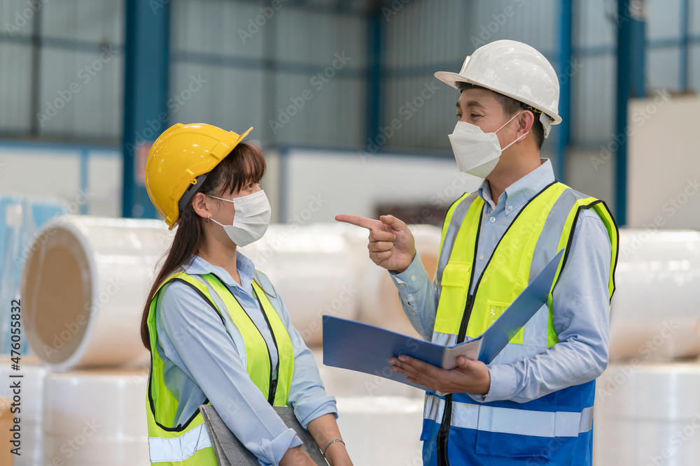 Asian male supervisor engineer in helmet safety wearing hygienic mask to protect coronavirus hold documents explaining and training with female technician at warehouse factory industrial Asian male supervisor engineer in helmet safety wearing hygienic mask to protect coronavirus hold documents explaining and training with female technician at warehouse factory industrial