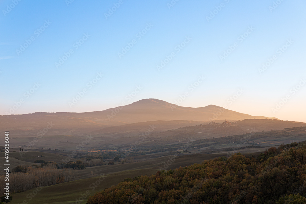 Photo of a Tuscan countryside panorama during sunset. In the foreground the cultivated fields and a wood, in the background some houses, the mountains and the sun's rays