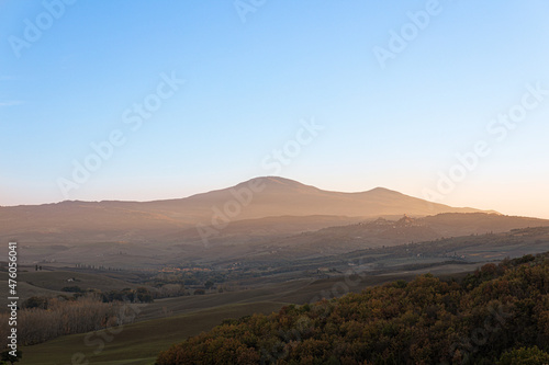 Photo of a Tuscan countryside panorama during sunset. In the foreground the cultivated fields and a wood, in the background some houses, the mountains and the sun's rays
