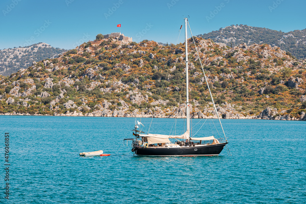 Fototapeta premium Cruise yacht sails on the sea near the sunken ancient town of Kekova in Turkey against the background of castle Simena on a high rock with Turkish flag