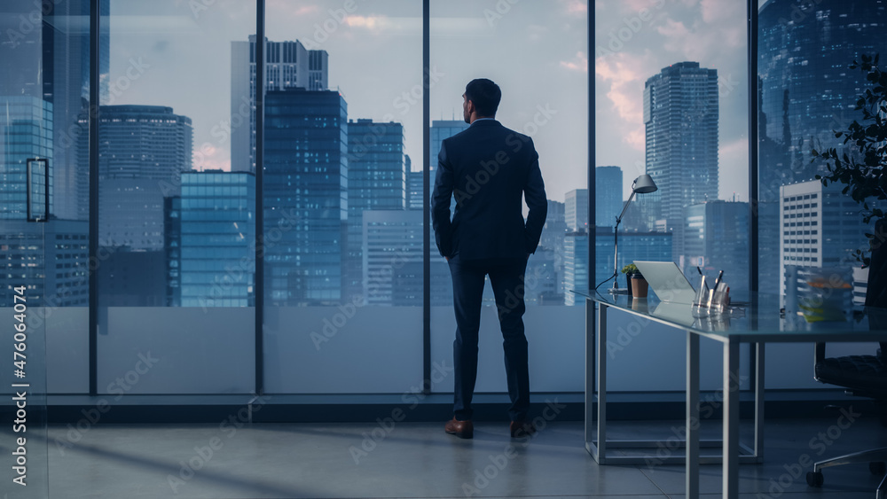 Confident Young Businessman in a Perfect Tailored Suit Standing in His Modern Office Looking out of the Window on Big City with Skyscrapers. Successful Finance Manager Planning Project Strategy.