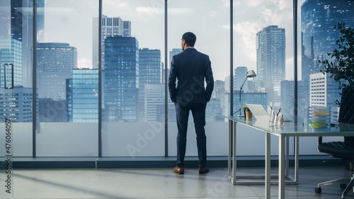Confident Young Businessman in a Perfect Tailored Suit Standing in His Modern Office Looking out of the Window on Big City with Skyscrapers. Successful Finance Manager Planning Project Strategy.