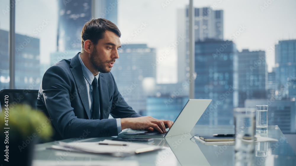 Happy Businessman in a Suit Sitting at a Desk in Modern Office, Using Laptop Computer, Next to Window with Big City with Skyscrapers View. Successful Finance Manager Planning Work Projects. Happy Businessman in a Suit Sitting at a Desk in Modern Office, Using Laptop Computer, Next to Window with Big City with Skyscrapers View. Successful Finance Manager Planning Work Projects.